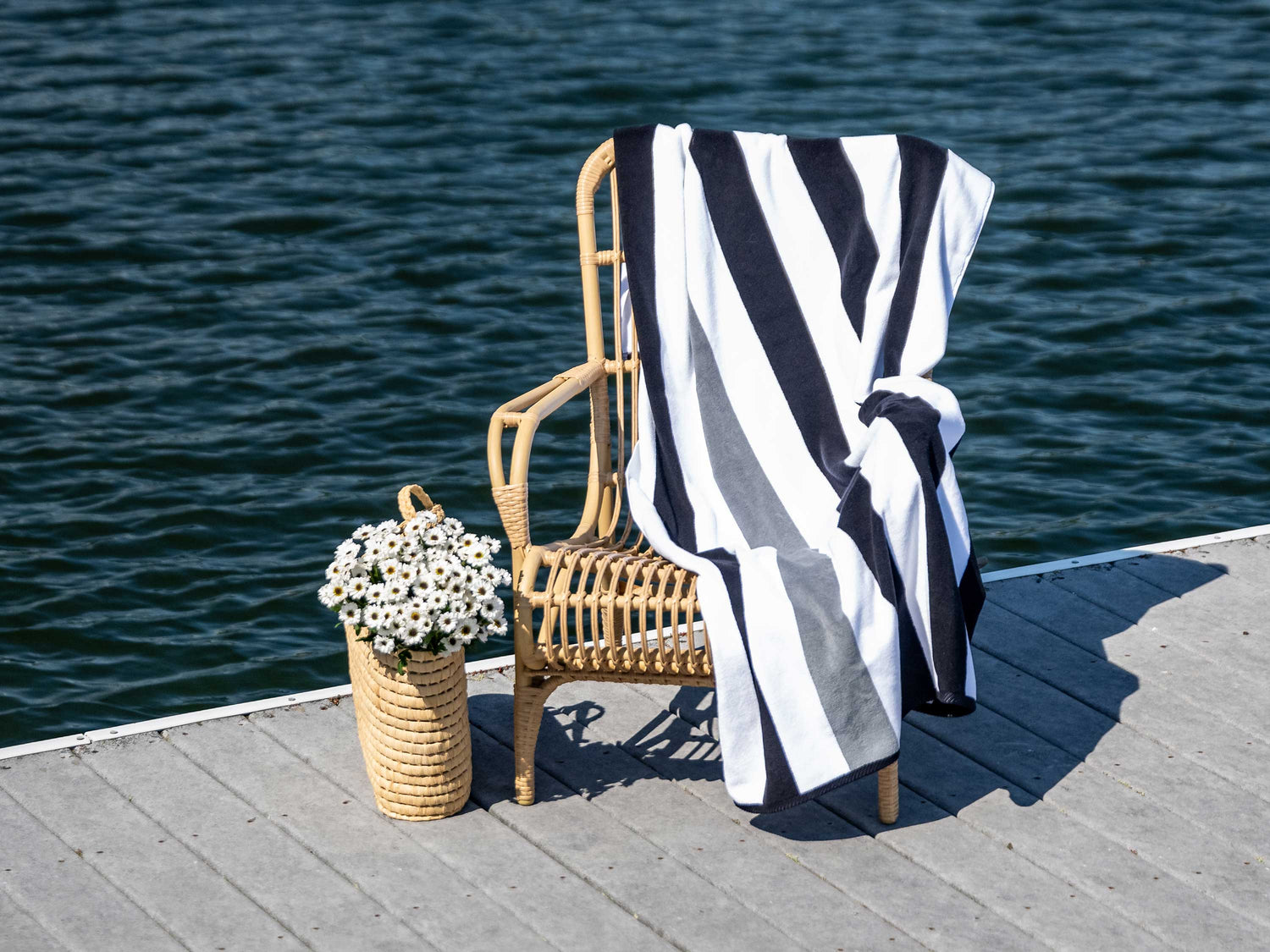 An extra-large, black, gray, and white striped cabana beach towel laid out on a chair next to a bag of flowers on a dock on a lake.