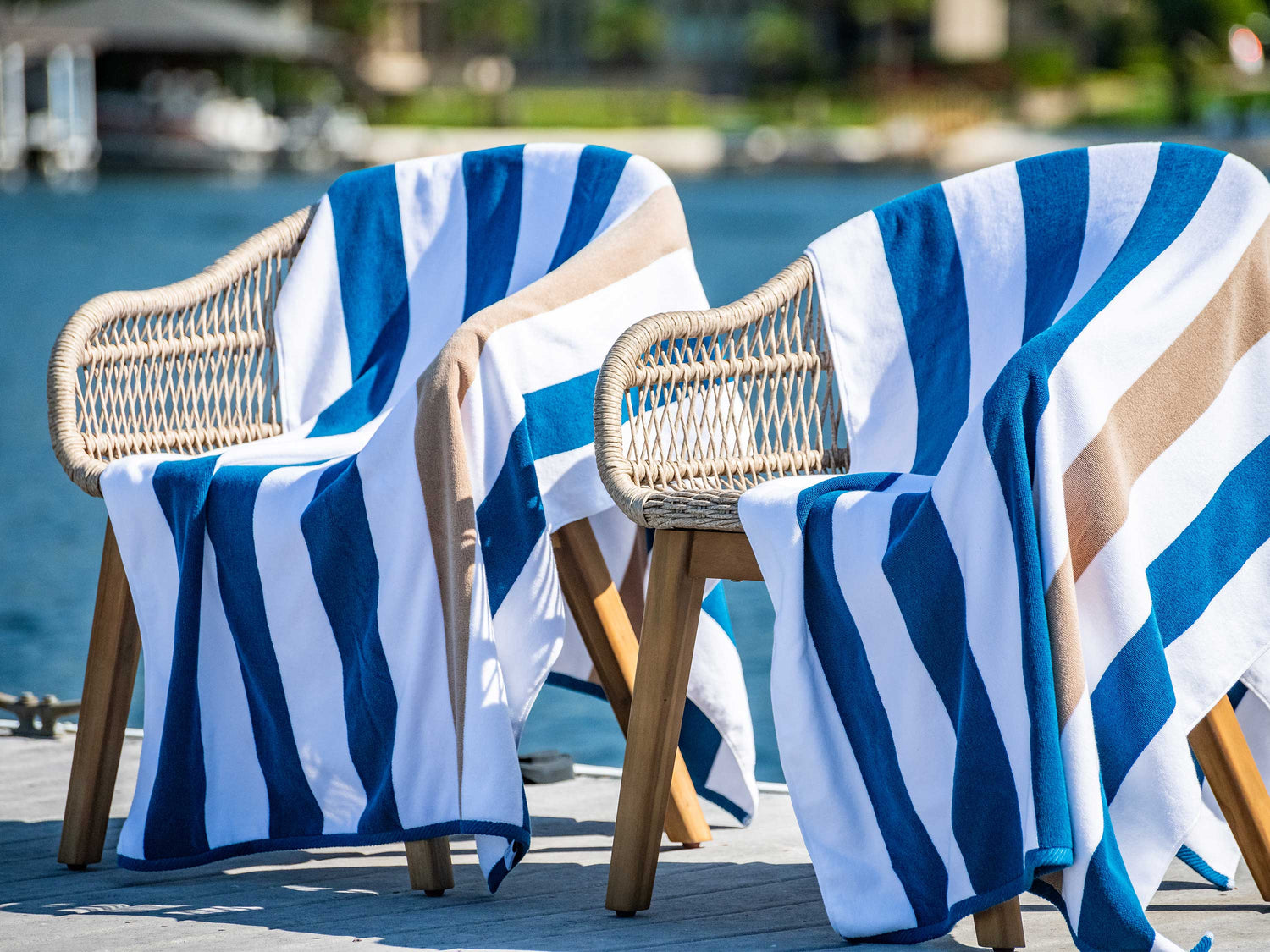 Navy blue, tan, and white striped cabana beach towels on wicker chairs on a dock on a lake.