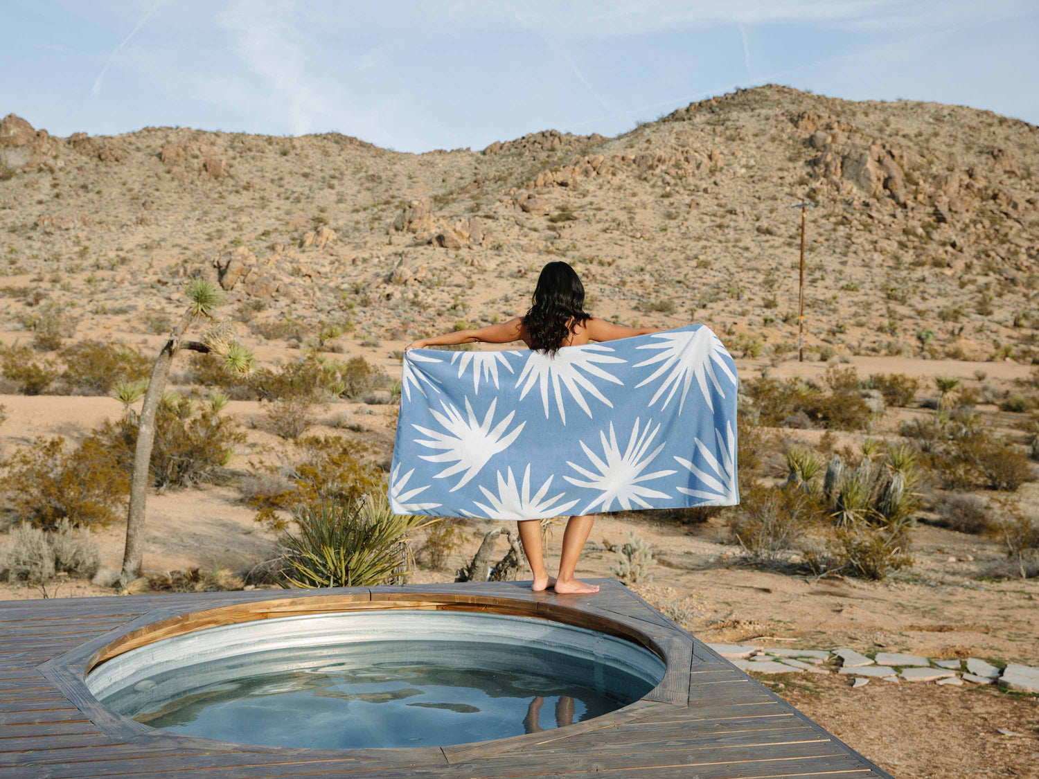 
      A woman standing beside a jacuzzi in the desert with her back turned, holding out a blue and white tropical patterned beach towel.
    