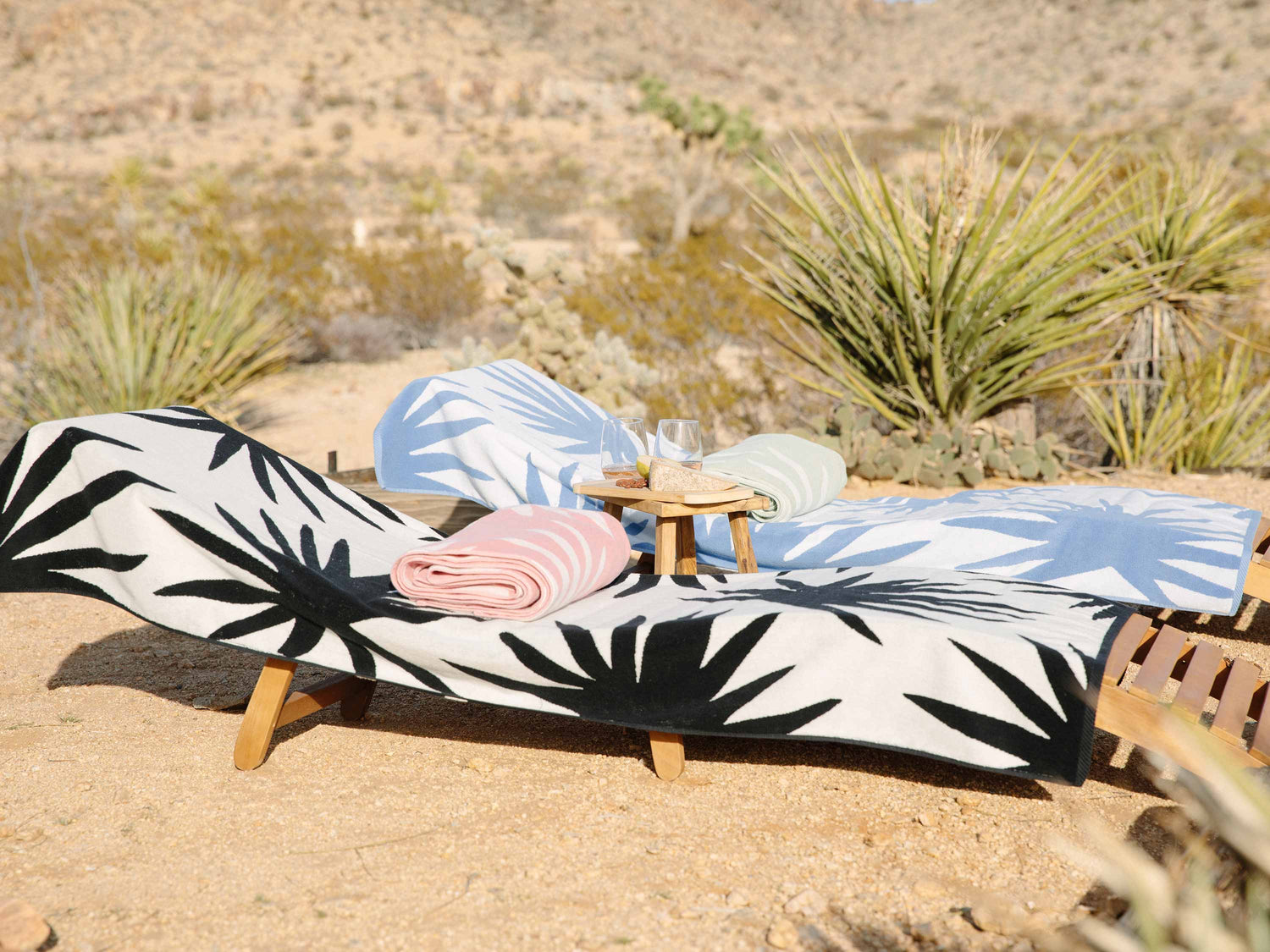 Tropical patterned beach towels laid out on two lounge chairs with a folded towel atop each and a small table with glasses in between.