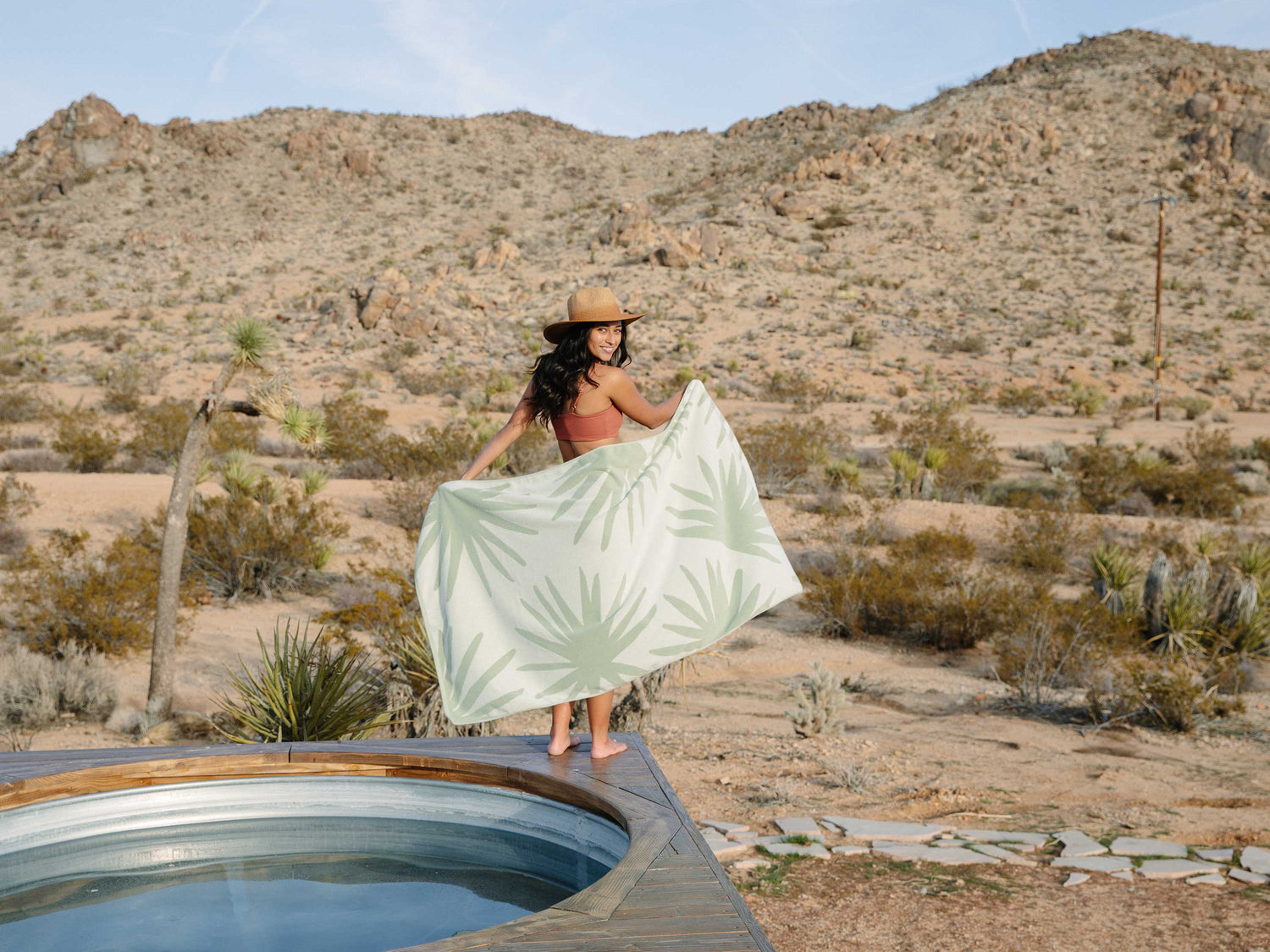 A woman wearing a hat looking backward, standing beside a jacuzzi in the desert and holding out a white and green tropical patterned beach towel.