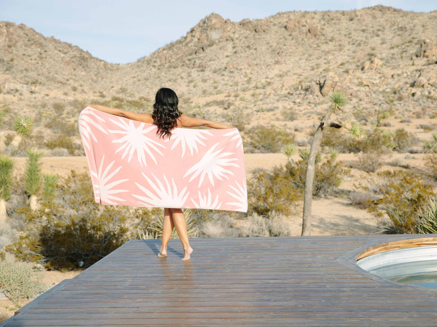 
      A woman standing on a deck in the desert with her back turned, holding out a pink and white tropical patterned cabana beach towel.
    
