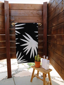 A folded black and ivory white palm tree patterned cabana beach towel hanging on a towel rack in an outdoor shower.