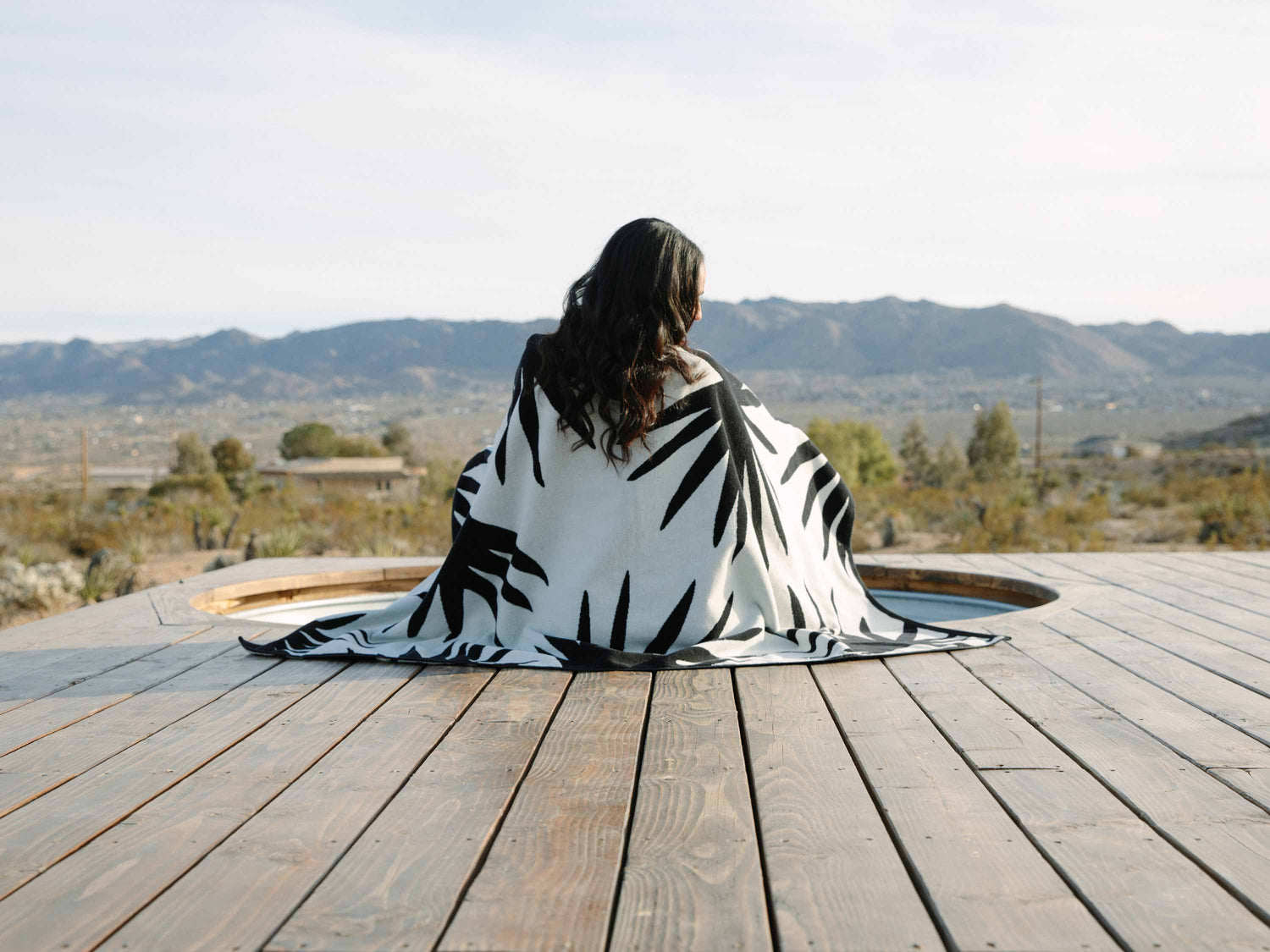 A woman sitting with her back turned and her feet in a jacuzzi, wrapped in a black and white tropical patterned cabana beach towel.