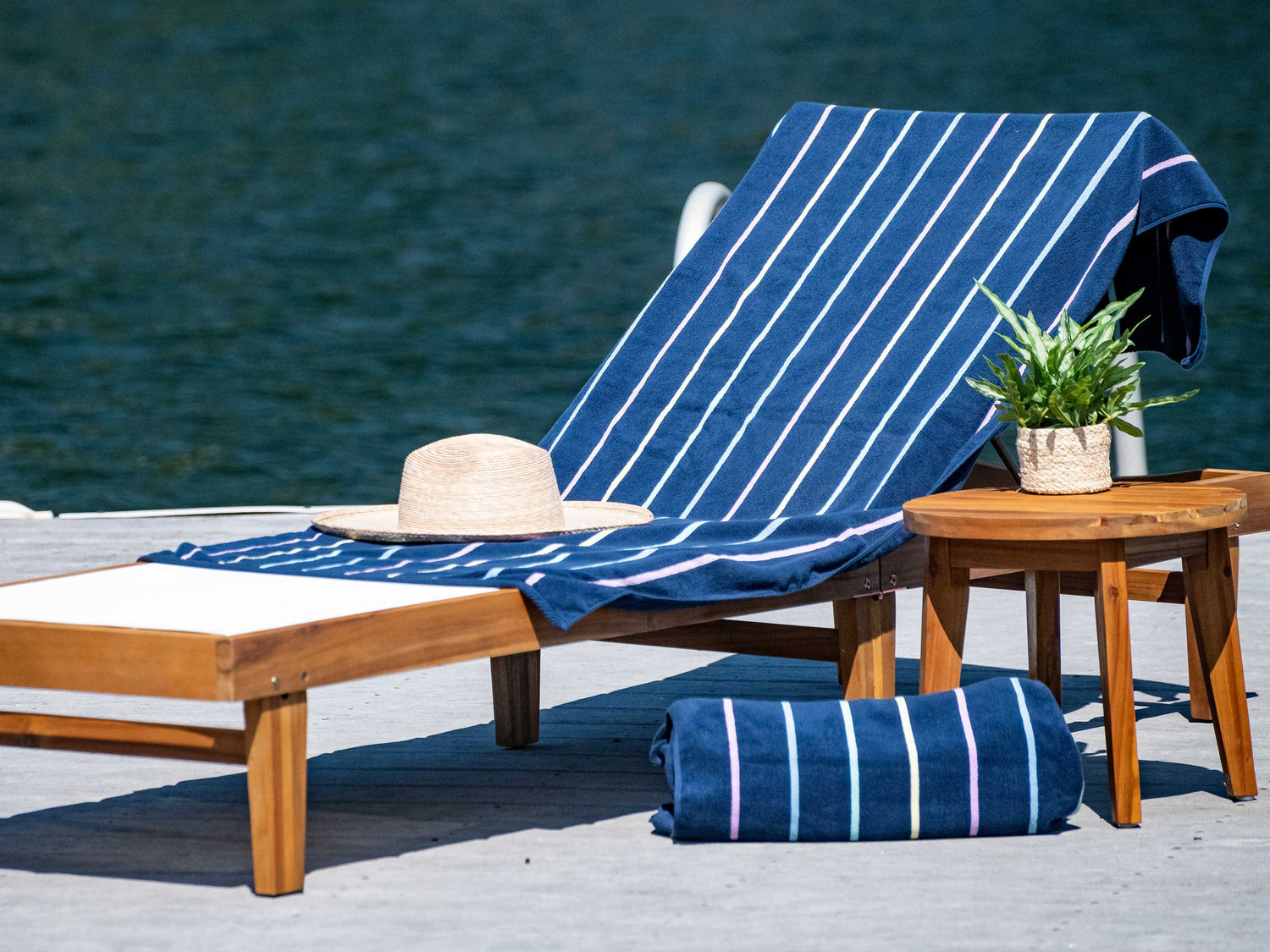A navy blue, striped towel laid out on a lounge chair beside a plant atop a small table and a rolled-up towel with a lake in the background.
