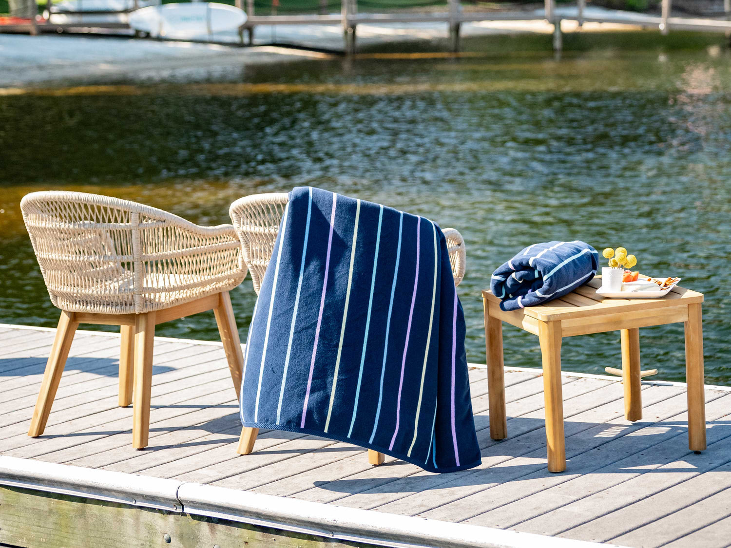 A navy blue, striped towel hanging over a chair in between another chair and a table with flowers and a rolled-up towel atop it on a dock.