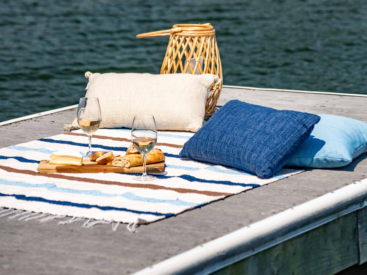 A picnic including wine and cheese on top of a white ivory, blue, and brown striped Mexican Blanket by the water.