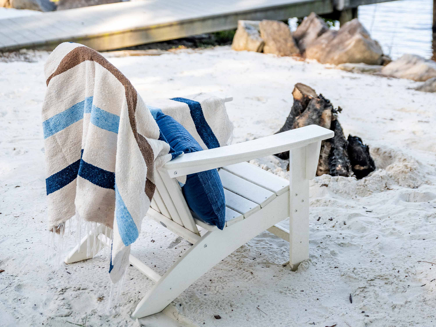 A white ivory, brown, and blue striped Mexican Blanket draped over a white chair on the sand.