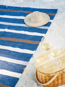 A picnic basket next to a navy blue striped Mexican Blanket on the sand.
