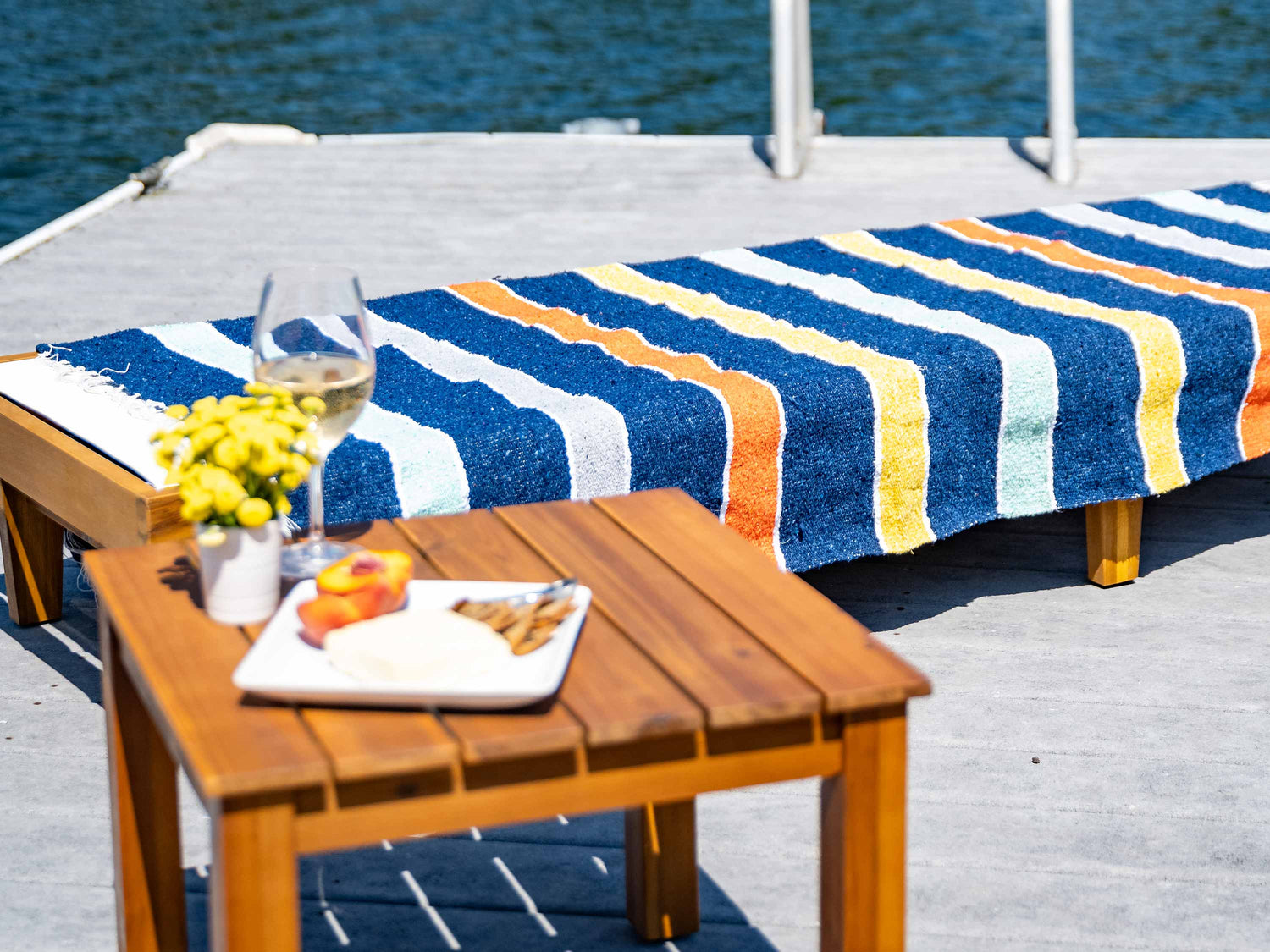 A lounge chair next to the water with a multicolored striped Mexican Blanket laid out on top of it.