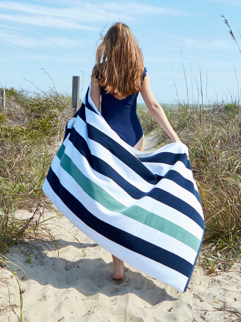 A woman walking on the beach while holding out a blue, green, and white striped cabana beach towel spread out behind her.