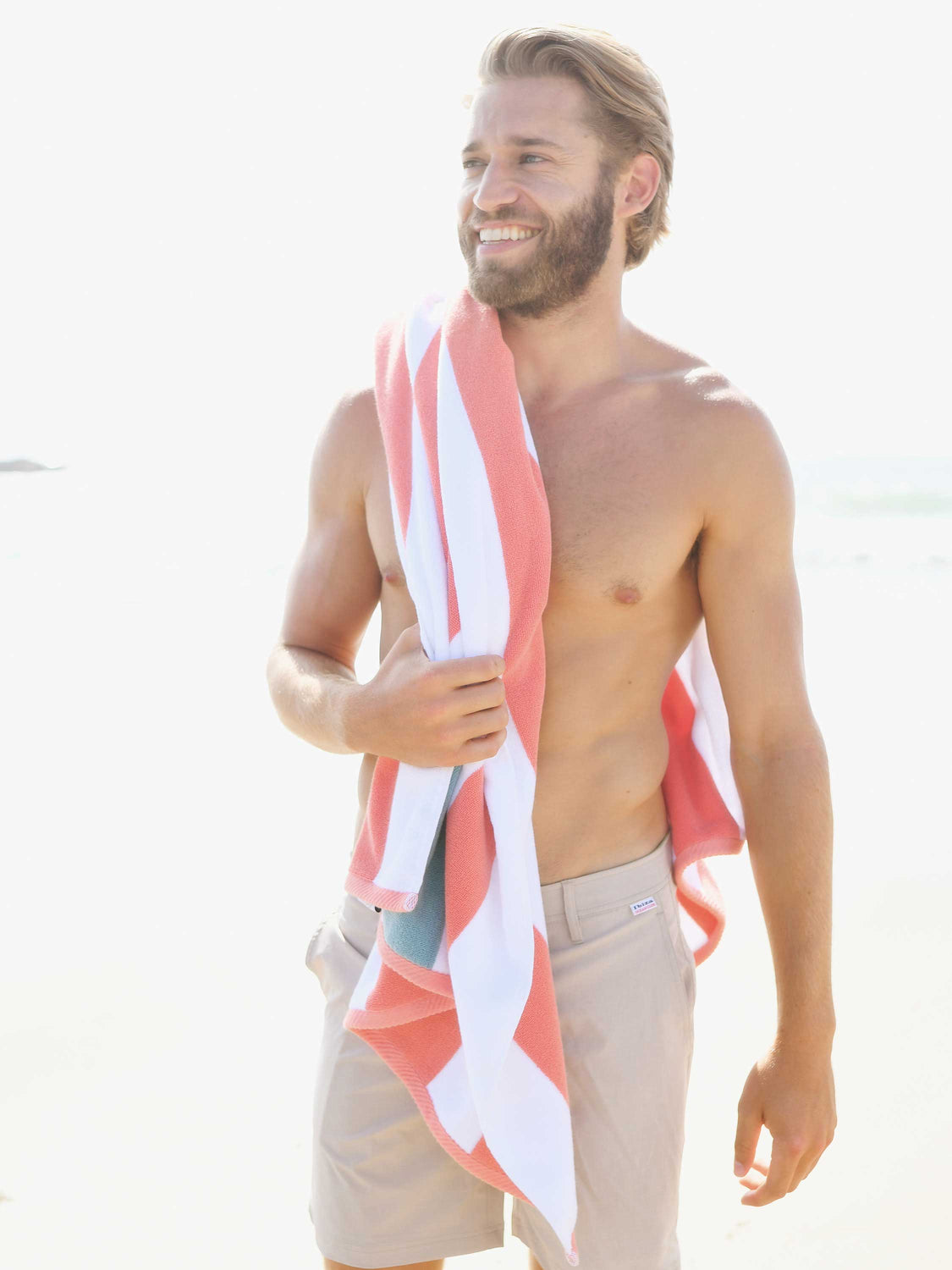 A man standing on the beach with an orange, green, and white striped cabana beach towel hanging over his shoulder.