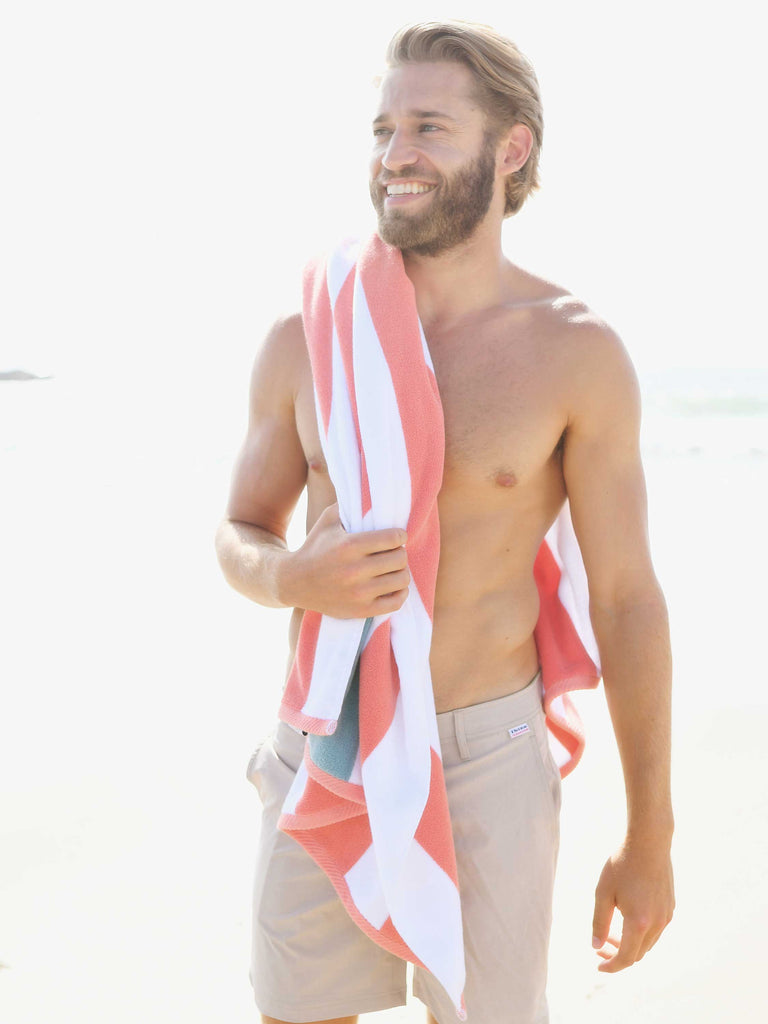 A man standing on the beach with an orange, green, and white striped cabana beach towel hanging over his shoulder.