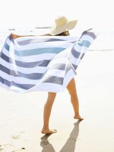 A woman walking on the beach while holding out a gray, green, and white striped cabana beach towel spread out behind her.
