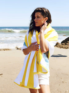 A woman standing on the beach with the ocean behind her, wrapped in a yellow, green, and white striped cabana beach towel.