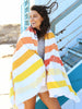 A woman on the beach wrapped in an extra large, yellow, orange, and white striped cabana beach towel.