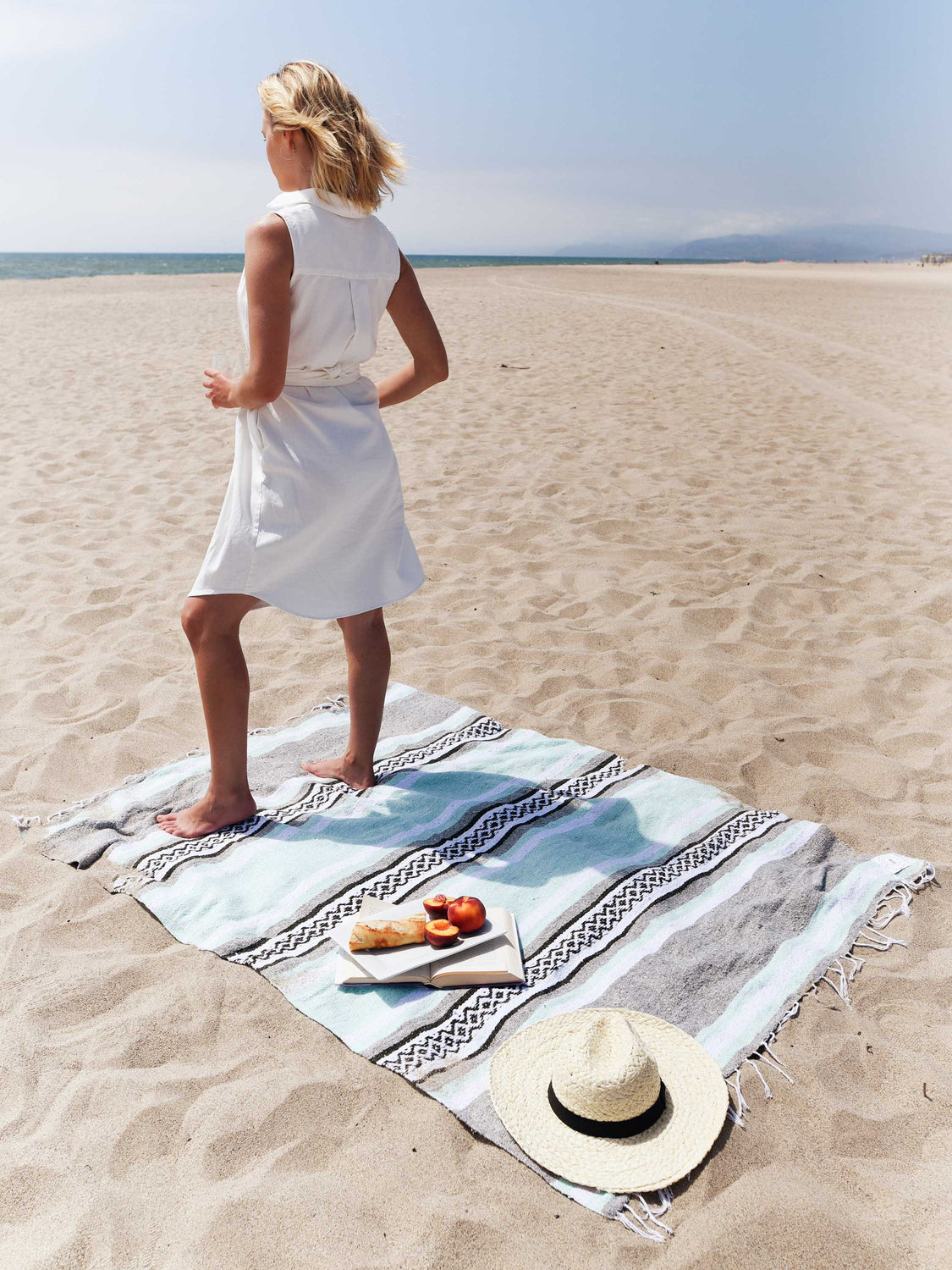 A woman standing on a gray and green Mexican blanket spread out on the beach with her hat, book, and fruit laid on top.