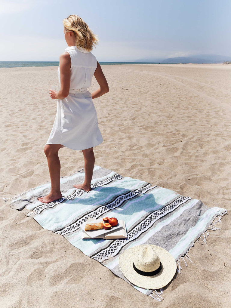 A woman standing on a gray and green Mexican blanket spread out on the beach with her hat, book, and fruit laid on top.