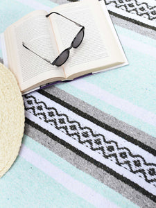 Close-up of an opened book and a pair of sunglasses on a gray, green, and white patterned Mexican blanket spread out. Blanket Selena Gomez was wrapped on on her Instagram Story. Selena Gomez Mexican Blanket.