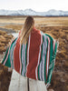 A woman standing on a marsh wrapped in an oversized green, red, and white striped Mexican blanket.
