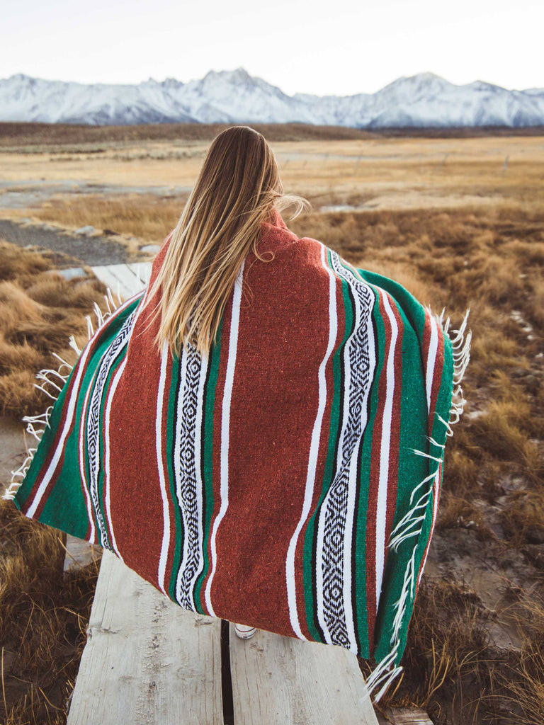 A woman standing on a marsh wrapped in an oversized green, red, and white striped Mexican blanket.