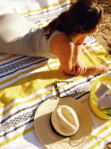 A woman lying outdoors on a yellow and white striped Mexican blanket with her hat, sunglasses, and bucket of drinks. 