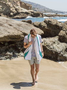 A man walking on the beach with an oversized aqua blue and white striped microfiber beach towel wrapped around his neck.