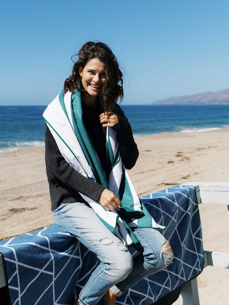 A woman sitting on a lifeguard tower railing with an oversized green striped microfiber beach towel wrapped around her neck.