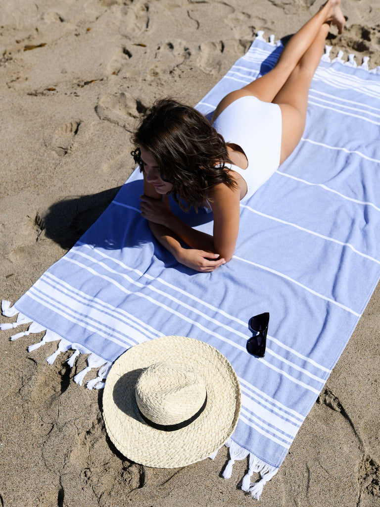 A woman in a white bathing suit laying out on an oversized blue and white striped Turkish towel at the beach.