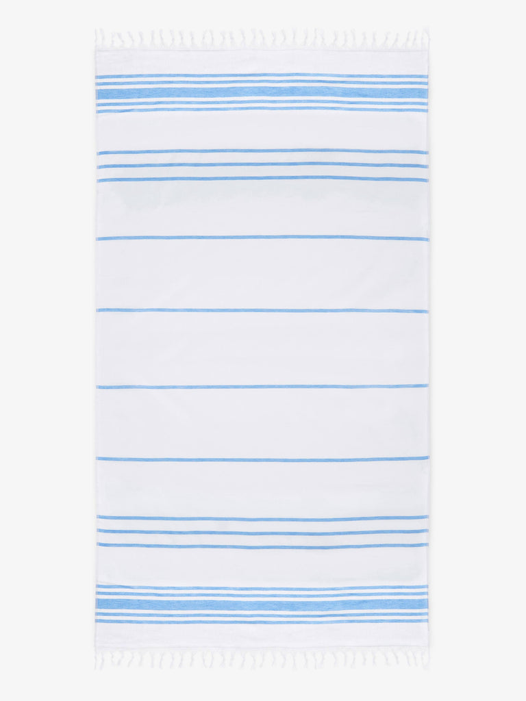 An oversized, white and light blue striped Turkish cotton towel with white fringe laid out.