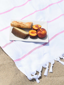 A charcuterie plate atop an oversized, white and pink striped Turkish beach towel laid out on the sand.