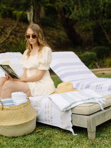 A woman reading a book outdoors, sitting on a lounge chair with a tan and white Turkish towel draped over it.