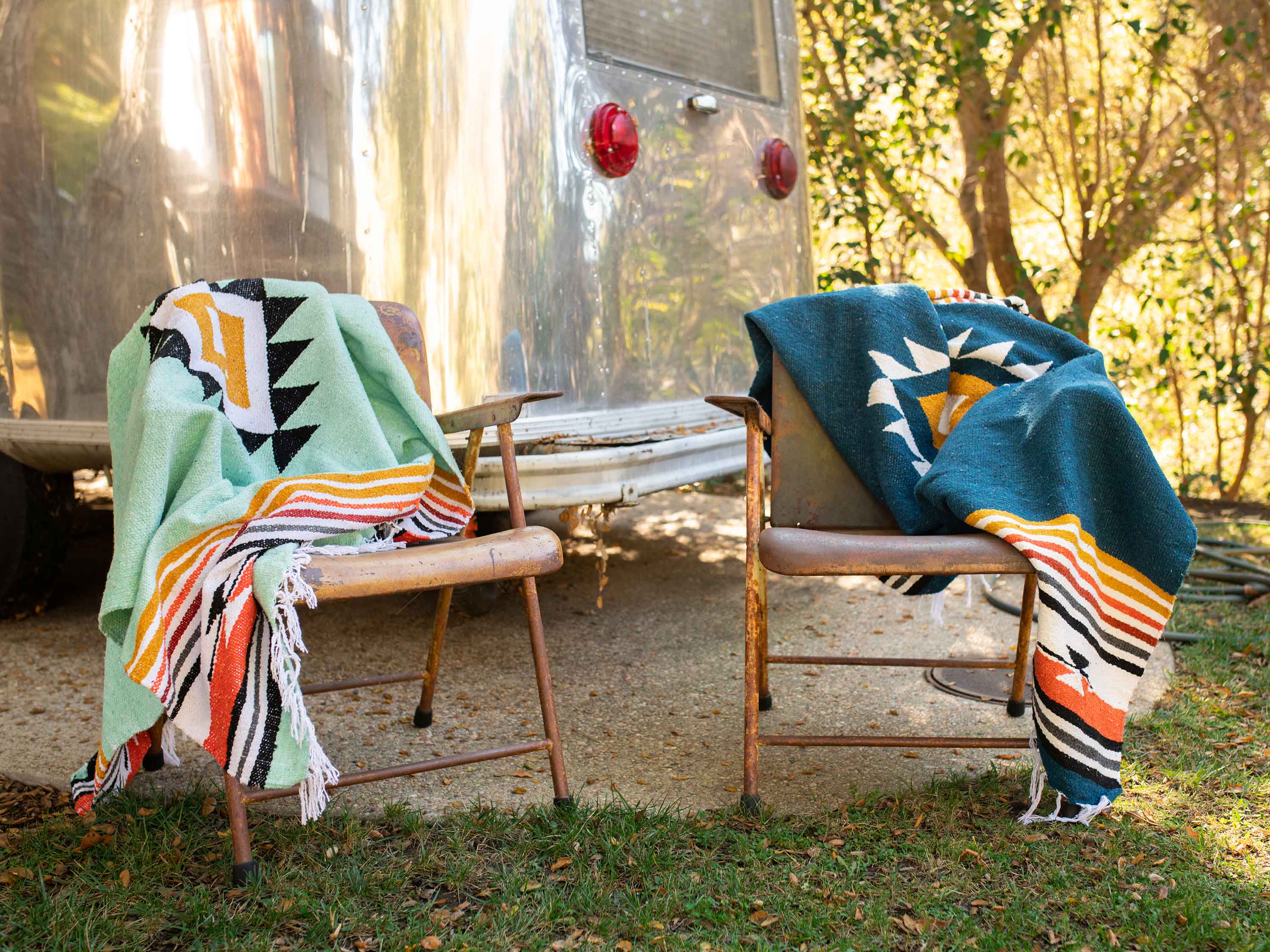 Teal and blue Mexican blankets draped on chairs outdoors, next to a silver camper. 