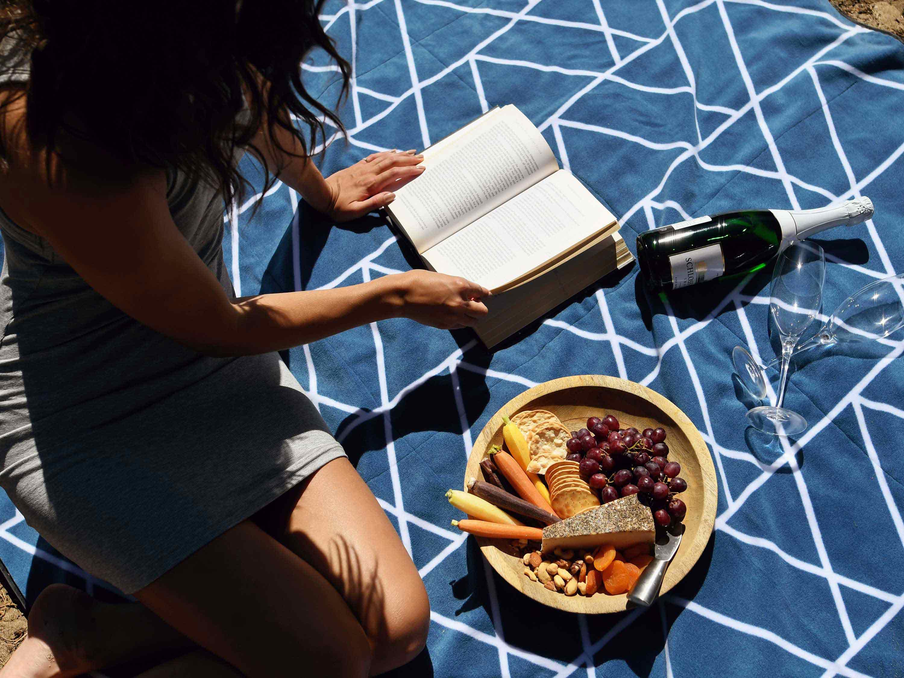 A woman sitting on a picnic blanket reading a book with a charcuterie board next to her.
