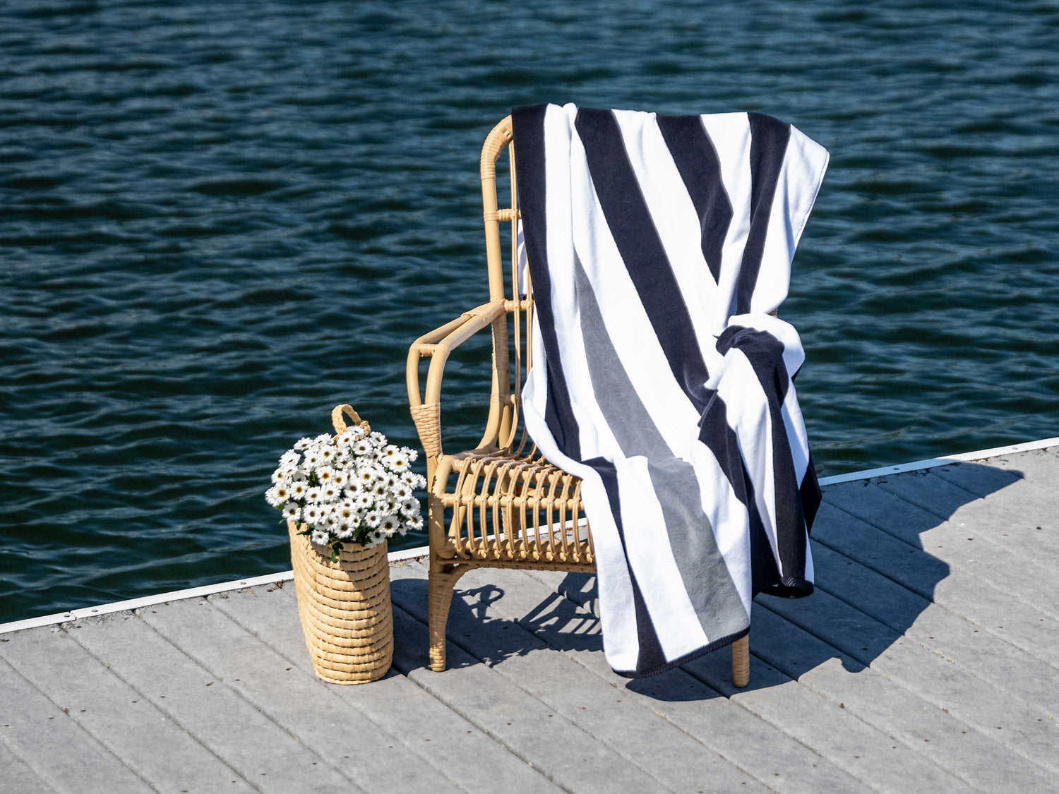 
      An extra-large, black, gray, and white striped cabana beach towel laid out on a chair next to a bag of flowers on a dock on a lake. 
    