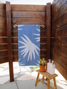 A folded blue and white palm tree patterned cabana beach towel hanging on a towel rack in an outdoor shower. 