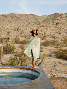 A woman wearing a hat, standing sideways beside a jacuzzi in the desert and wrapped in a green and white tropical patterned cabana beach towel.