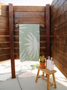 A folded green and white palm tree patterned cabana beach towel hanging on a towel rack in an outdoor shower. 