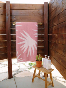 A folded pink and white palm tree patterned cabana beach towel hanging on a towel rack in an outdoor shower. 