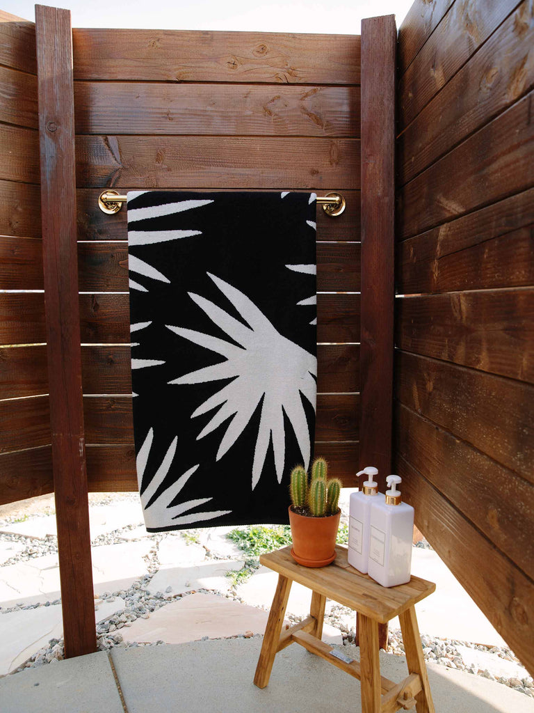 A folded black and ivory white palm tree patterned cabana beach towel hanging on a towel rack in an outdoor shower. 