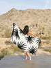 A woman standing on a deck in the desert, holding out a black and ivory white tropical patterned cabana beach towel spread out behind her.