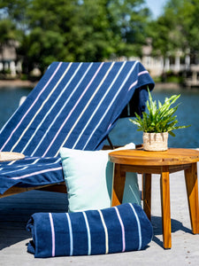 A close-up of a navy blue, striped towel laid out on a lounge chair beside a plant atop a small table, a green pillow and a rolled-up towel.