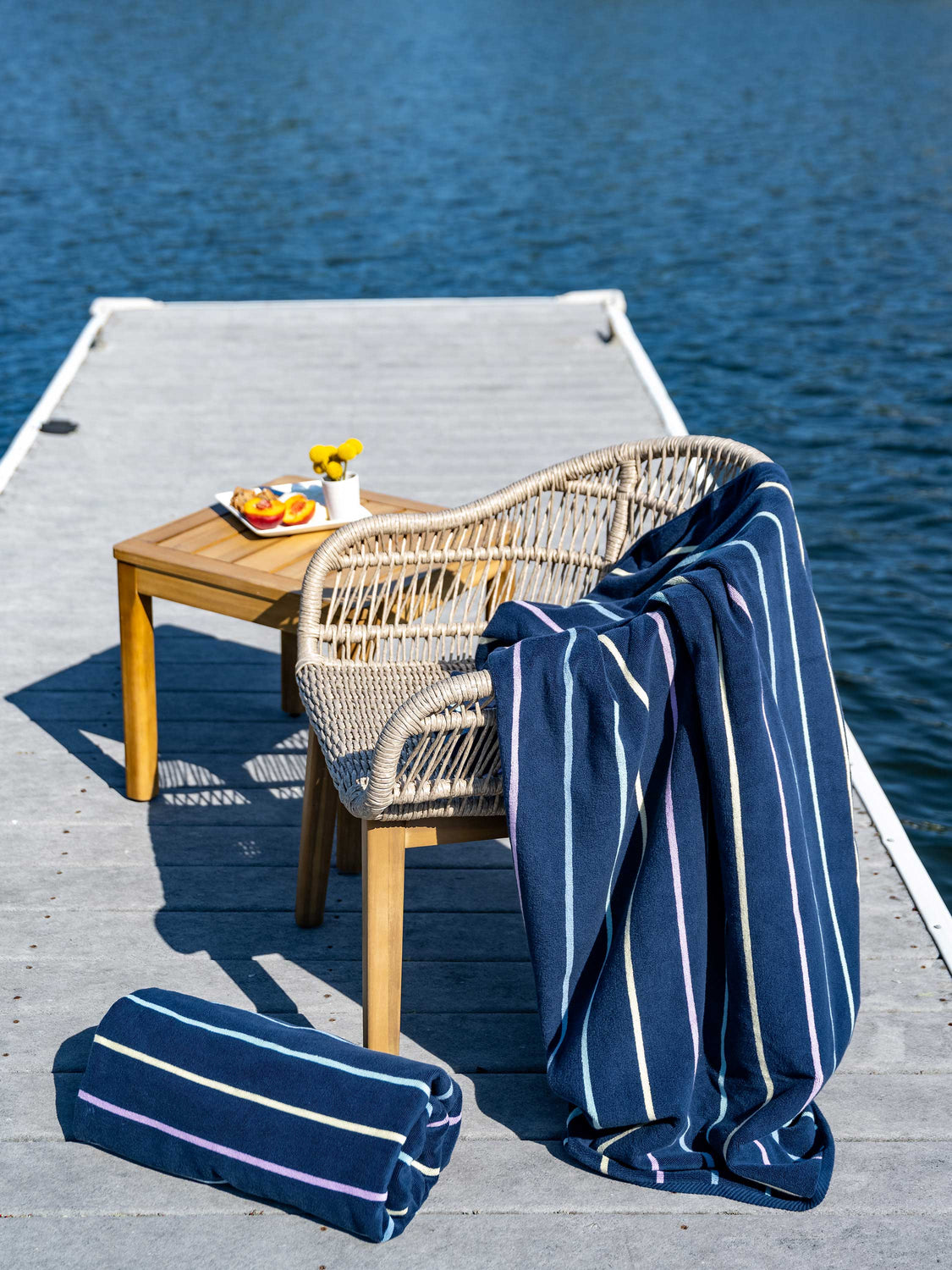 A navy blue, striped towel hanging over a chair in between a table with a plate of fruit and flowers atop it and a rolled-up towel on a dock.
