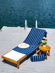 A navy blue, striped towel laid out on a lounge chair beside a plant atop a small table and a rolled-up towel with a lake in the background.