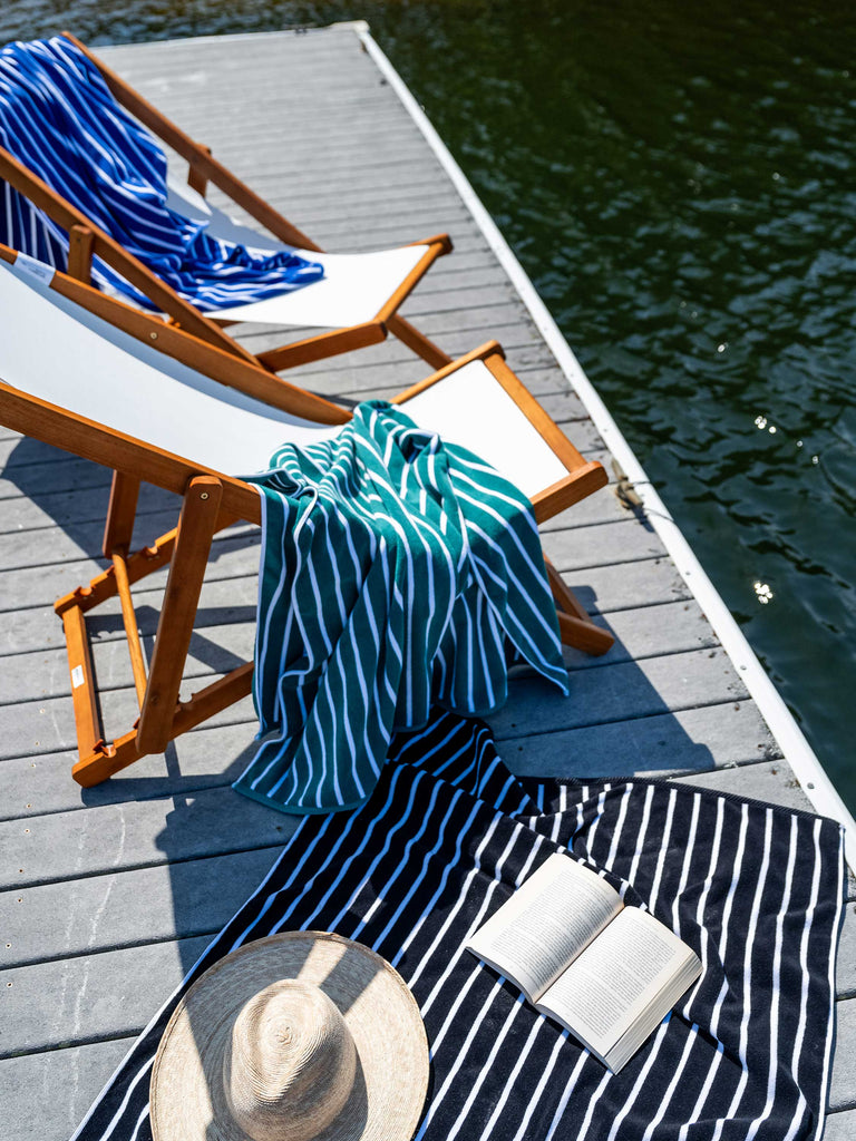 Blue, green, and black striped extra-large cabana towels draped on luxury deck chairs on a lake dock.