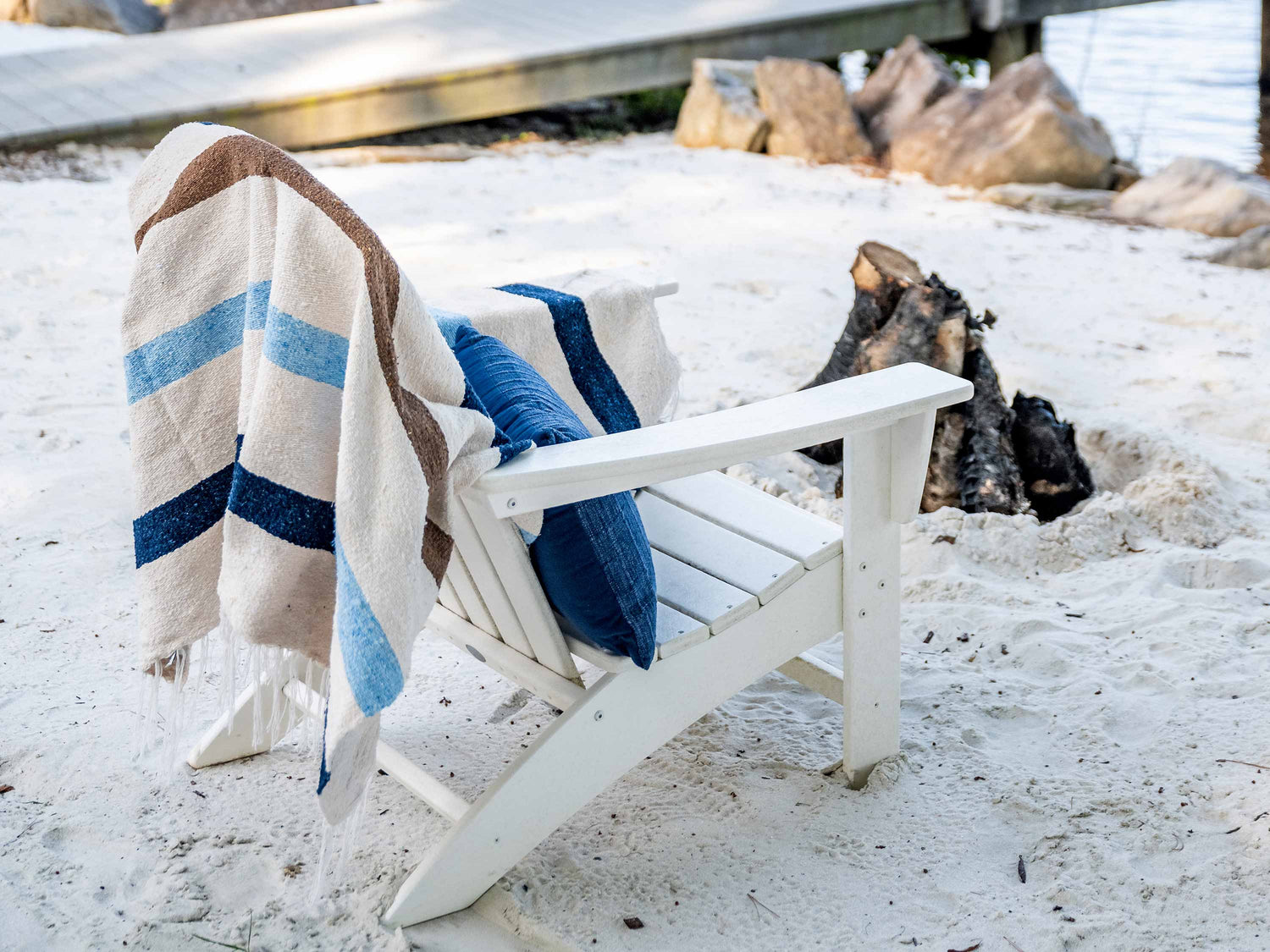 
      A white ivory, brown, and blue striped Mexican Blanket draped over a white chair on the sand.
    