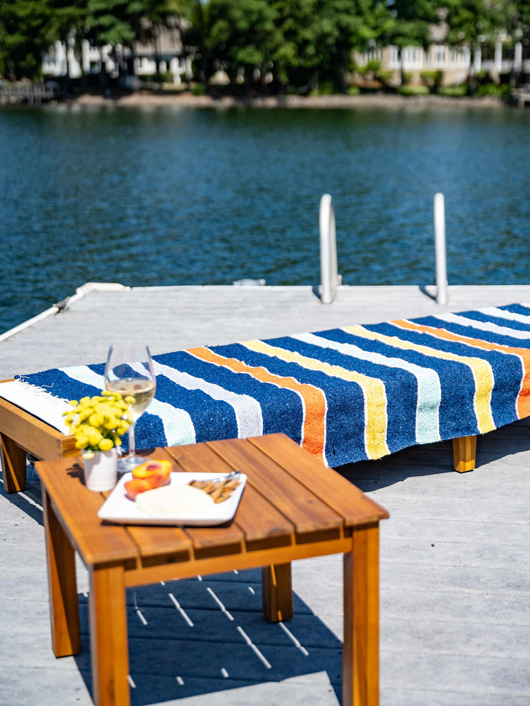 A lounge chair next to the water with a multicolored striped Mexican Blanket laid out on top of it.