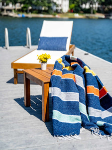A lounge chair next to the water with a multicolored Mexican Blanket in the foreground.