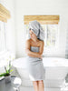 A woman sitting on the edge of a bathtub wrapped in a cloud gray bath towel with her hair also wrapped in a cloud gray bath towel.