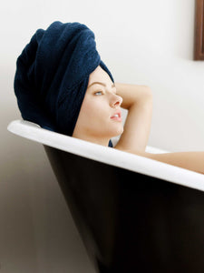 A woman laying in a bathtub with her hair wrapped in a navy blue cotton bath towel.
